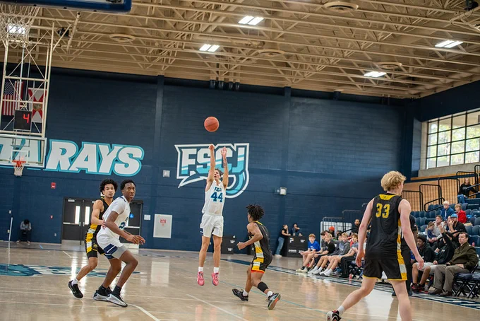 A basketball player in white jersey number 44 jumps to shoot the ball mid-air during a game in a gym with 'RAYS' and 'FSCJ' logos on the wall.