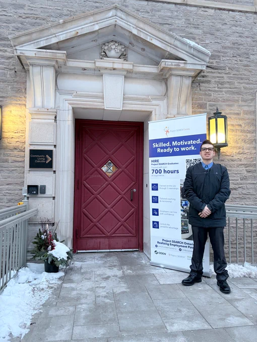 Christopher D'Ambriosia, Project SEARCH graduate, stands in front of the event venue with a display promoting Project SEARCH Canada