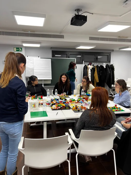 Otro plano general de la mesa con las piezas de lego, al rededor están las alumnas cogiendo piezas para colocar en su tapete y hablando entre ellas.