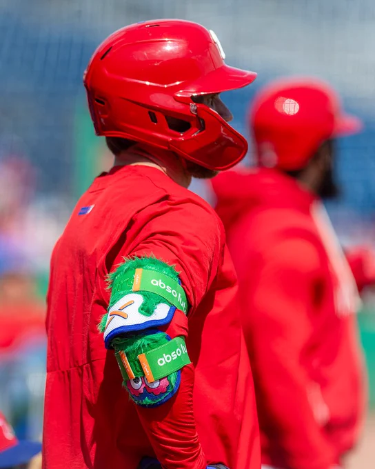 Bryce Harper waits to take batting practice at Phillies Spring Training. He wears a red Phillies workout shirt and helmet. On his elbow is a green Phillie Phanatic-inspired elbow guard.