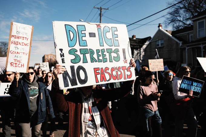 A crowd marches down a residential street in daylight while a protester in the foreground holds a large sign reading “DE-ICE THE STREETS / NO FASCISTS.” Other signs in the crowd include “Stand with Minnesota” and a poster that reads, “The only minority ruining this country are the rich.”