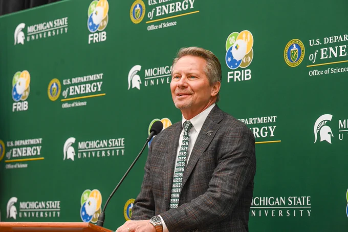 A man in a grey suit stands before a green backdrop with logos of Michigan State University, U.S. Department of Energy, and FRIB. He wears a tie with 'S' patterns and speaks at a podium with a microphone.