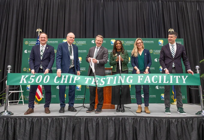 Six individuals formally cut a green ribbon that reads \"K500 CHIP TESTING FACILITY.\" Backdrops bear logos of the U.S. Department of Energy and Michigan State University. A person in the middle right is holding oversized scissors.