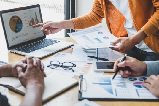 Three professionals gathered at a table looking at printed materials and charts on a laptop. 