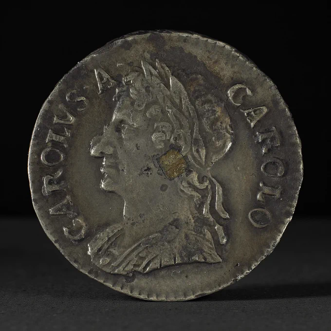 Pièce de métal gris montrant un profil d’homme avec une couronne de laurier et un petit carré de métal foncé près de l’oreille.