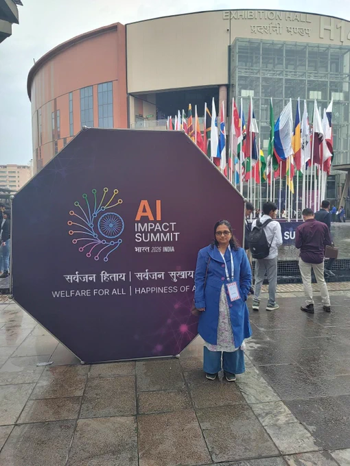 A person stands in front of a large “AI Impact Summit Bharat 2026 India” display outside an exhibition hall, with multiple international flags in the background. The person is wearing a blue coat and conference badge, standing on a wet pavement, indicating recent rain. The summit slogan “Welfare for All | Happiness of All” is visible on the display.