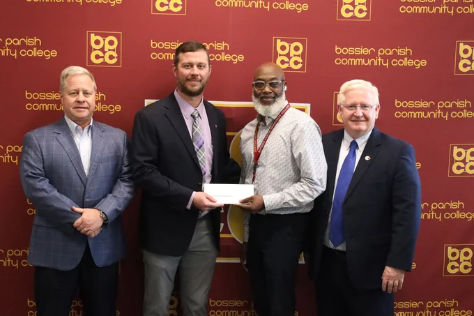 Four individuals are standing in front of a backdrop featuring the Bossier Parish Community College logo. One person is holding an envelope. They are dressed in business attire and smiling.