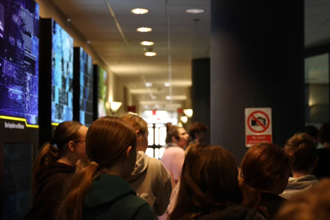 People standing in a dimly lit hallway with illuminated digital displays on the left wall.