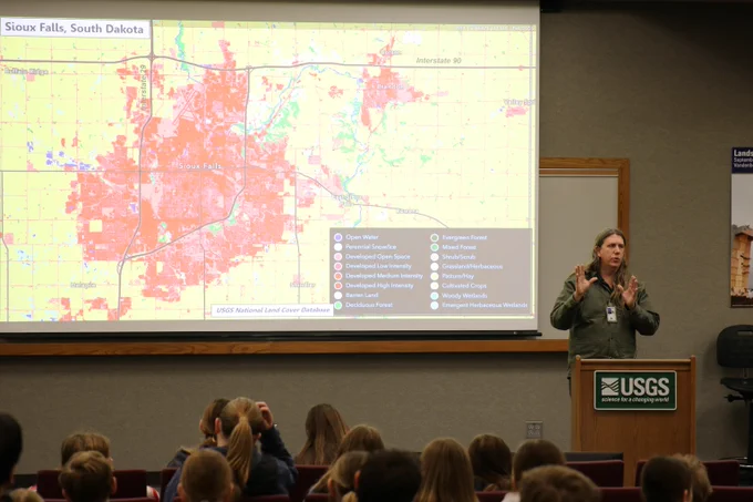 Presenter at USGS podium explaining a detailed map of Sioux Falls, South Dakota, to an attentive audience in a lecture room.