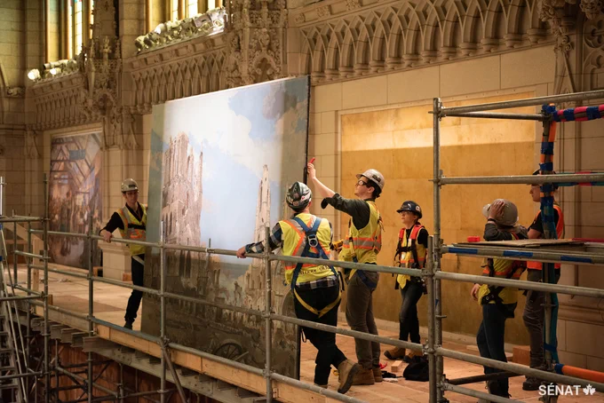 Cinq travailleurs se tiennent sur un échafaudage à l’intérieur de l’édifice du Centre, retirant avec soin une grande peinture historique.