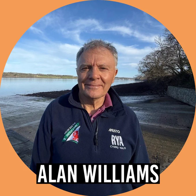 Alan Williams standing outdoors by a waterfront on a sunny day, wearing a navy RYA Cymru-Wales jacket, with calm water and trees in the background.