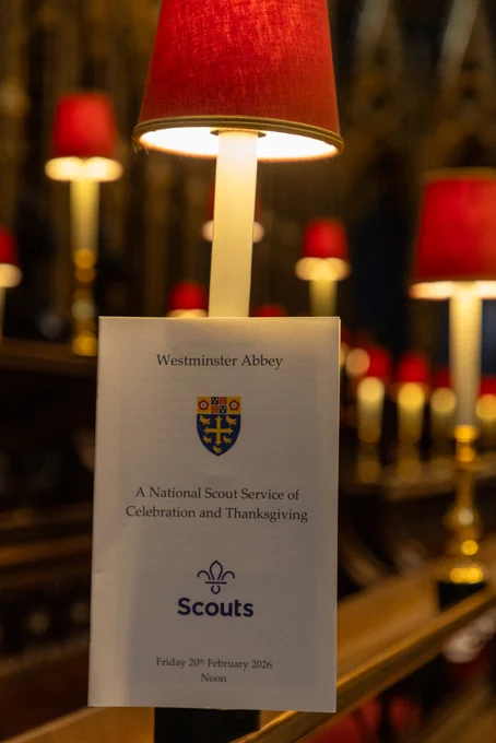 An order of service is placed on a pew beneath a lamp with a red shade at Westminster Abbey. The front cover features a coat of arms and reads: 

Westminster Abbey
A National Scout Service of Celebration and Thanksgiving
Friday 20th February 2026,

In the background, more lit lamps with red shades are visible, adding to the warm ambiance.