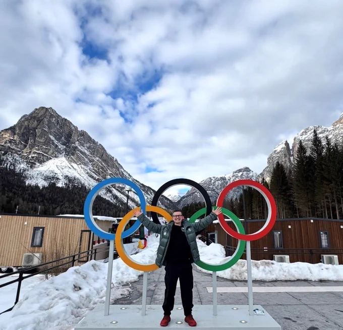Alt text: A person stands smiling with arms raised in front of large Olympic rings, with snow-covered ground and rocky mountain peaks in the background under a cloudy sky.