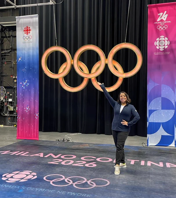 Alt text: A smiling person stands on a studio floor branded “Milano Cortina 2026,” posing with one arm raised toward large illuminated Olympic rings, flanked by CBC and Milano Cortina banners on a stage set.