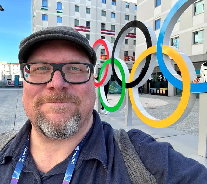 Alt text: A smiling person takes a selfie in front of large colourful Olympic rings in an outdoor plaza, with buildings in the background.