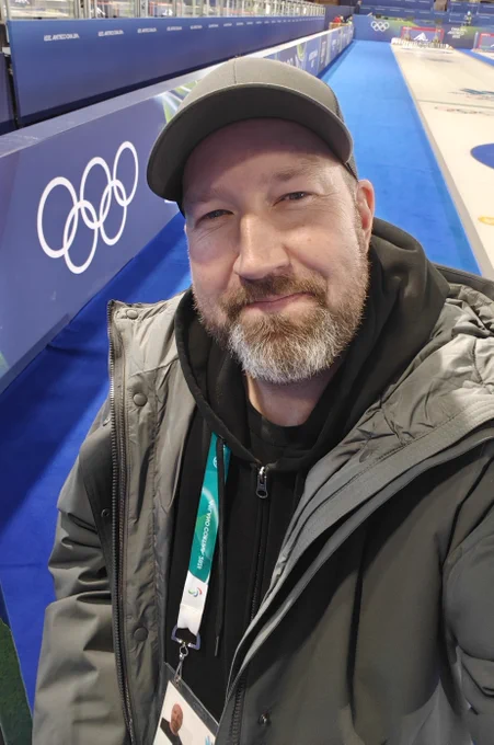 Alt text: A person takes a selfie beside an Olympic curling rink, with the Olympic rings visible on the boards and blue ice surface in the background.