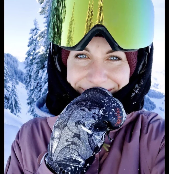 Eine wunderschöne Frau mit einem leichten Lächeln in Wintersachen mit Handschuhen, Mütze und Skibrille. Im Hintergrund sieht man eine Winterlandschaft.