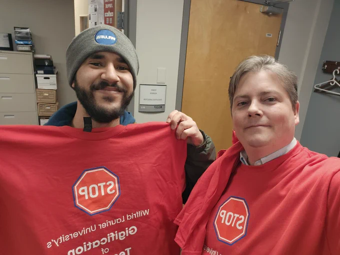 Ryan Ladner and a Laurier grad student wearing and showing the new, red, Contract Faculty solidarity shirt. Grad student is also wearing a WLUFA toque.