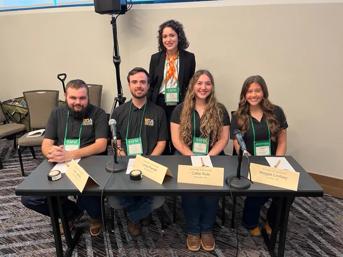 A group of five individuals at a panel table with microphones. Four are seated with name cards reading: Sam Jones, Linus Parrish, Callie Rule, and Maggie Lindsey. The standing person is behind the seated group. They are wearing name badges and smiling.