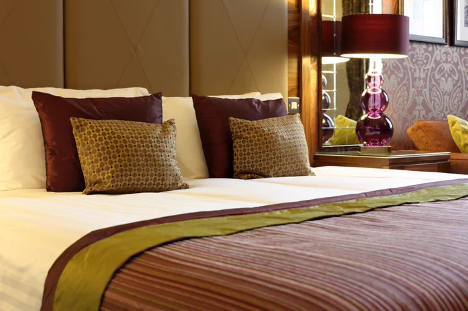 Luxurious hotel room featuring a neatly made bed with decorative pillows, a stylish lamp, and elegant wallpaper.