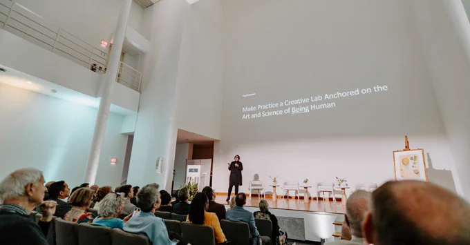 A person stands on a stage in front of an audience in a modern auditorium. The screen behind displays the text "Make Practice a Creative Lab Anchored on the Art and Science of Being Human." The audience is seated and attentive.