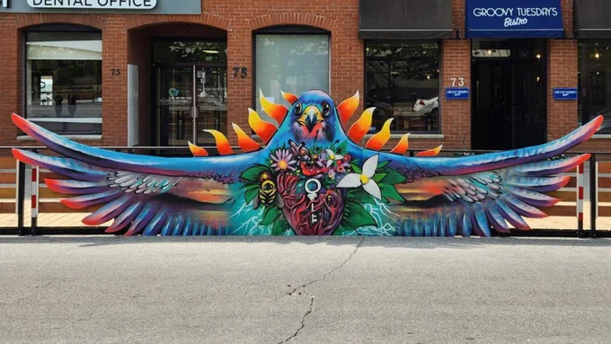 A colorful mural painted on a sidewalk barrier depicts a vibrant bird with outstretched wings. The bird is embellished with a heart at its center featuring various symbols and flowers. A brick building in the background has "Groovy Tuesdays Bistro" and "Dental Office" signs.