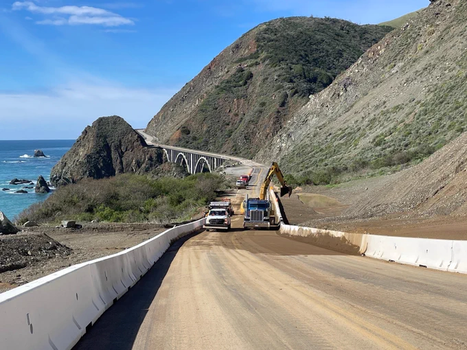 Picture is from road level looking south on Hwy. 1 at Regent's Slide showing crews and equipment clearing debris from the roadway and catchment area. Big Creek Bridge is in background.