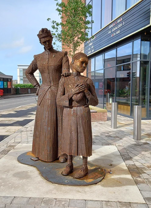 Rust-coloured metal statue of a Victorian woman standing sternly behind a young girl holding a loaf of bread, commemorating Sophia Constable, the youngest female inmate at HM Prison Northallerton, displayed outside a modern building.