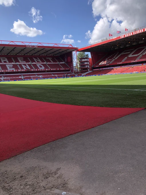 Photo taken inside Nottingham Forest’s City Ground stadium. The shot is taken in daylight at pitch/ground level. In the immediate foreground to the left is a red carpet whist to the right is a gravel track. The slightly shaded, green pitch can be seen in the centre of the image with the adjoining Trent End and Brian Clough stands meeting diagonally towards the back of the image, these stands are completely empty. At the very top of the photo blue sky dominates the left hand side whilst the right is shrouded in thick white, fluffy cloud.