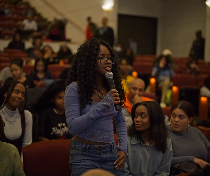 Young woman is standing in an auditorium holding a microphone during a discussion or Q&A session. Photo: Marshall Wayne Cooper