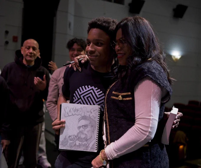 Young person holding a sketchbook with with a drawing of Malcolm X. He standing next to Dr. Ilyasha Shabazz. The two are posing for a photo. Photo: Marshall Wayne Cooper