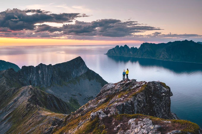 Two hikers standing on a mountain peak overlooking the ocean at sunset with rugged cliffs in the background.