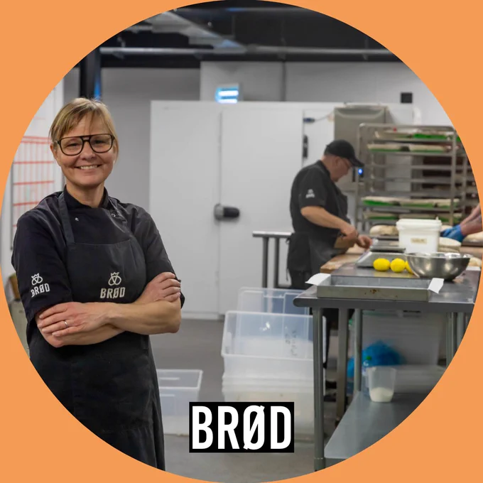 A woman in a BRØD uniform stands smiling in a commercial kitchen, while another staff member works at a counter in the background.