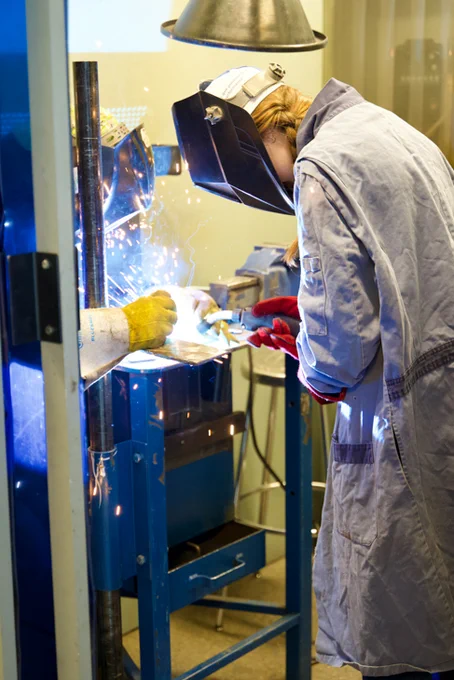 A person wearing protective gear is welding metal with bright sparks visible. They are in a shop classroom setting with a blue welding machine. Another person assists by steadying the metal with a gloved hand.