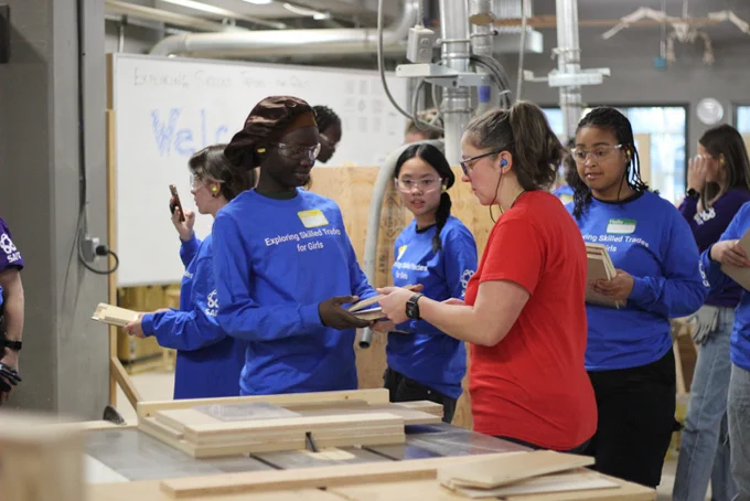 A group of people in a workshop setting, some wearing shirts with "Exploring Skilled Trades" and holding wooden items. One person in a red shirt is handing a piece of wood to another individual. In the background, there are various tools and equipment.