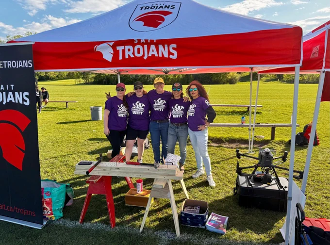 Five people stand together smiling under a red and white tent labelled "SAIT Trojans" at an outdoor event. They are wearing matching purple Women in Trades and Technology shirts. The area includes a grassy field, a wooden table with various items, and a small equipment case nearby.