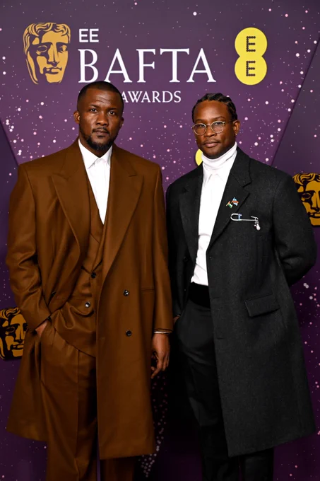 Wale Davies and Akinola Davis Jr. pose on the EE BAFTA Red Carpet. Wale is wearing a brown suit and Akinola is wearing a black suit.