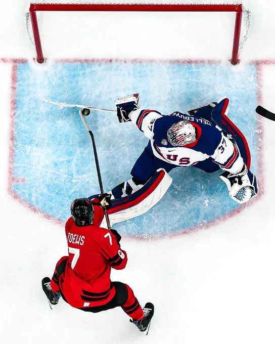 Photo of Connor Hellebuyck in a USA uniform making a save on Devon Toews in a Canada uniform