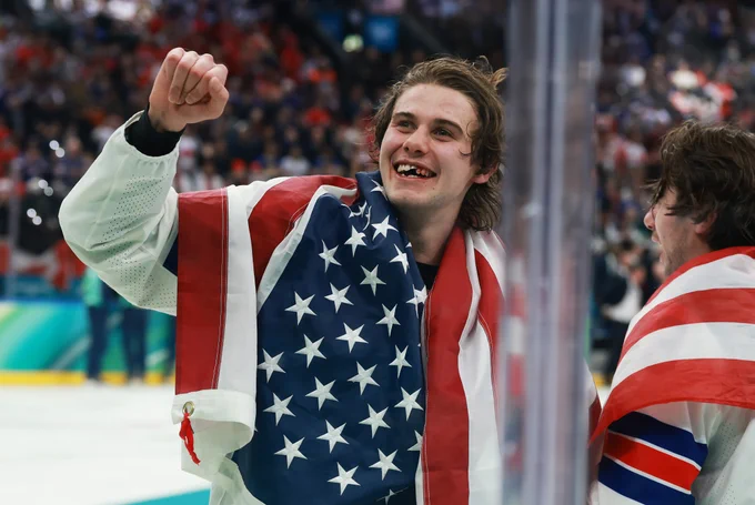 Jack Hughes on ice in a USA uniform with an American flag draped around his neck. He is holding his fist up and smiling showing a chipped tooth.