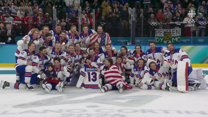 Group of men in Team U.S.A. hockey gear with gold medals around their neck taking a picture on the ice, with a jersey in front that reads "Gaudreau 13".
