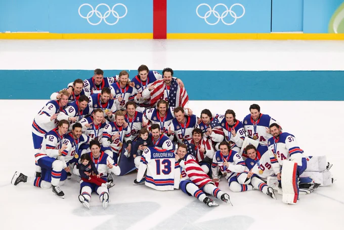 A group of ice hockey players in USA jerseys are joyfully posing on the ice with a flag draped over some of them. They're in a celebratory pose with Olympic rings visible in the background.