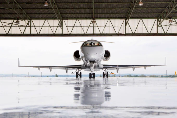 Frontal view of an aircraft parked in a hangar, with a glossy runway reflecting the plane and the hangar structure.