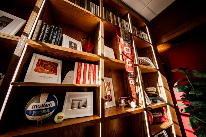 A well-organized wooden bookshelf filled with various items, including books, framed photos, a Molten-branded NCAA volleyball, and a University of Oklahoma penant. A potted plant is visible to the right. The shelves are illuminated, highlighting the diverse collection.
