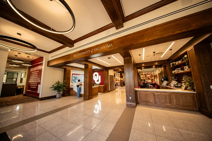 A spacious hall at the University of Oklahoma, featuring a reception area with a prominent university logo. The space is well-lit with elegant lighting fixtures, wooden beams, and a tiled floor. Decorative shelves with various items are visible, and a person stands near a wall display on the left.