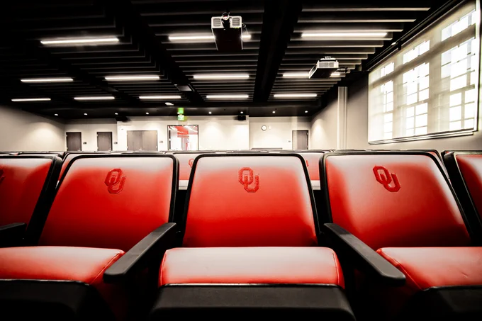 Red auditorium seats with the University of Oklahoma logo embroidered on the backrests are arranged in rows. The room features a modern design with black ceiling panels and large windows allowing natural light. A projector is mounted on the ceiling.