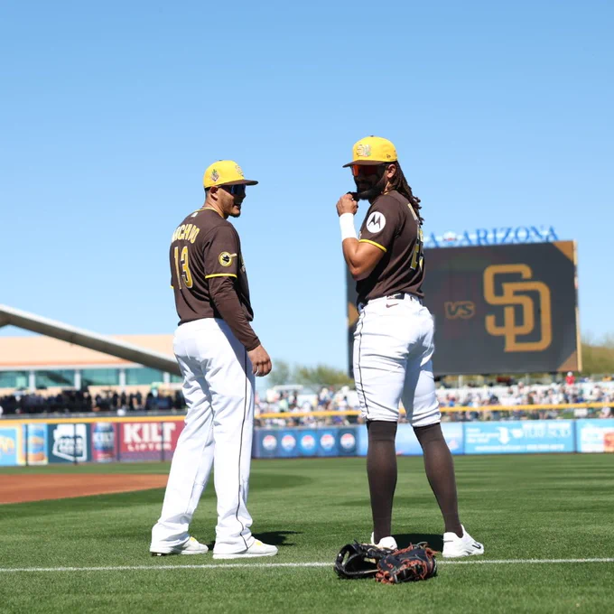 Manny Machado and Fernando Tatis Jr. look backwards at the camera. They wear brown Padres jerseys and yellow Spring Training caps. (Photo: Brendon Baranov/MLB)