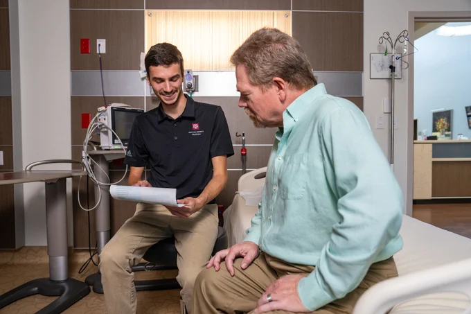 A person in a black shirt is sitting in a hospital room, talking to another person in a light green shirt who is sitting on a hospital bed. The person in black is holding a paper and smiling. Medical equipment is visible in the background.