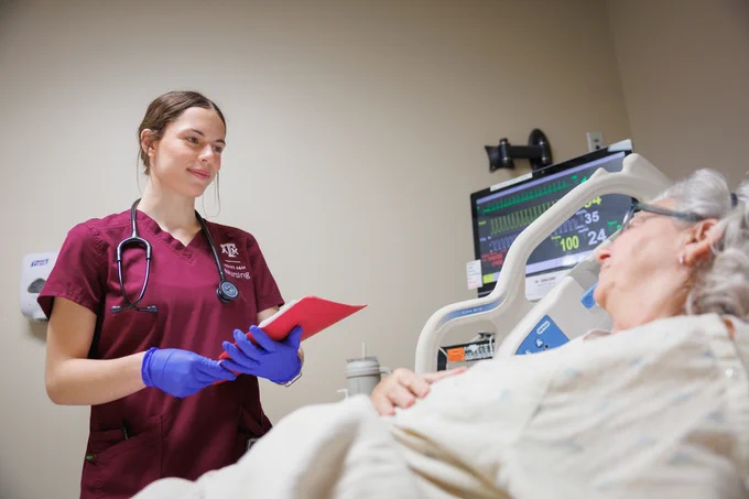 A healthcare worker is standing next to a hospital bed, holding a red clipboard and wearing a stethoscope. The person in the bed is resting. A monitor displaying vital signs is visible in the background.
