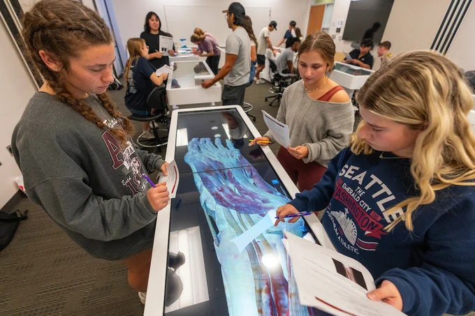 A group of students in a classroom is gathered around a digital anatomy table displaying a human foot. They are focusing on the image while holding papers. One student is wearing a sweatshirt with "Seattle Washington" printed on it. Other tables with students can be seen in the background.