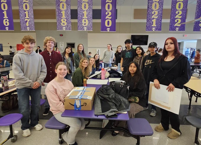 A large group of approximately 15 high school students poses for a group photo in a brightly lit school cafeteria.  In the foreground, a girl in a pink sweatshirt sits at a purple table with an Amazon cardboard box taped shut in front of her. Other students stand or sit around the long table, which is scattered with art supplies, bags, and personal items.  The students are dressed in casual clothing like hoodies, jeans, and sweaters. Hanging from the ceiling above them are large purple banners featuring various years (2016 through 2022) in gold lettering. The background shows the expansive cafeteria space with more tables and a few other people in the distance.